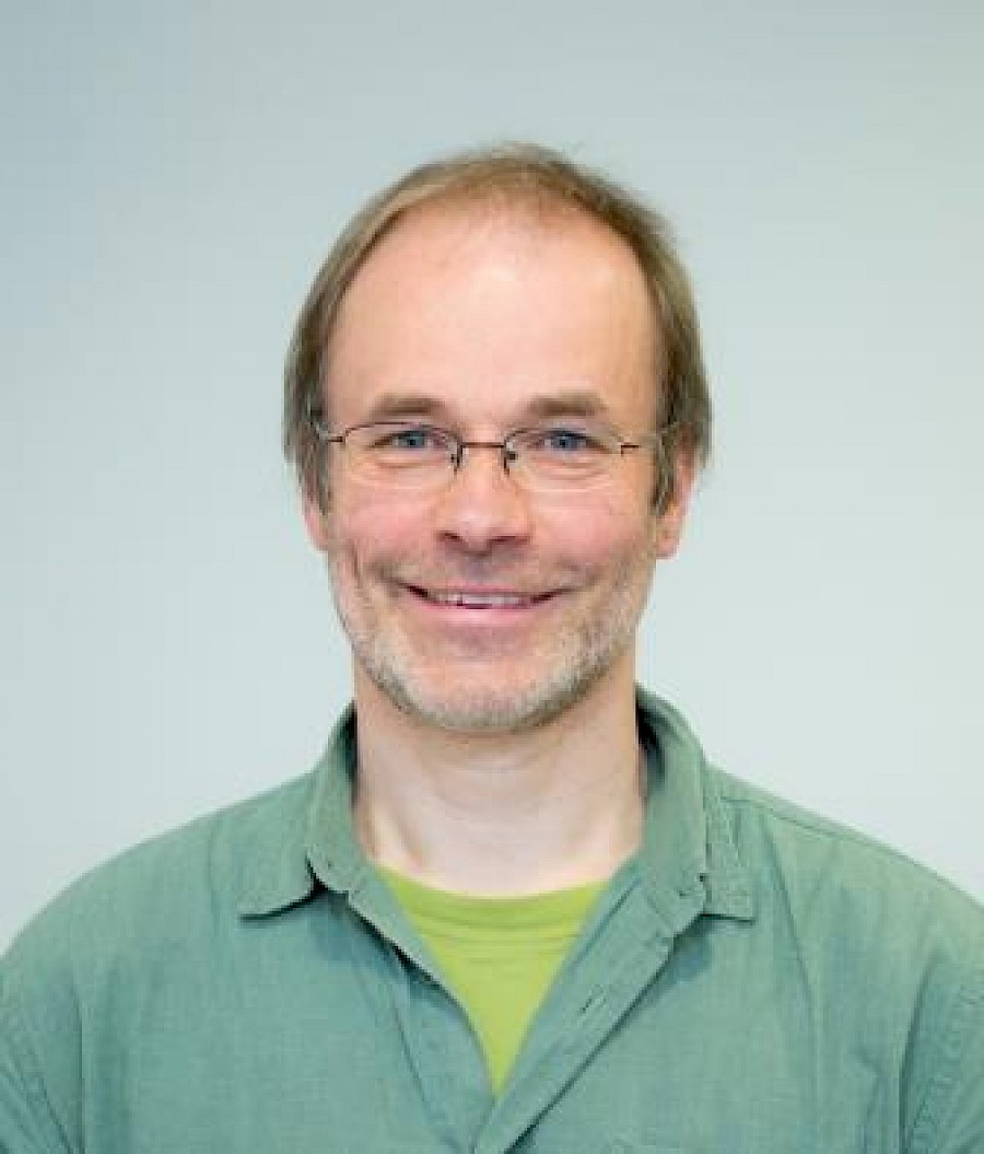 Eine männlich gelesene Person mit grünem Hemd und Brille lächelt in die Kamera